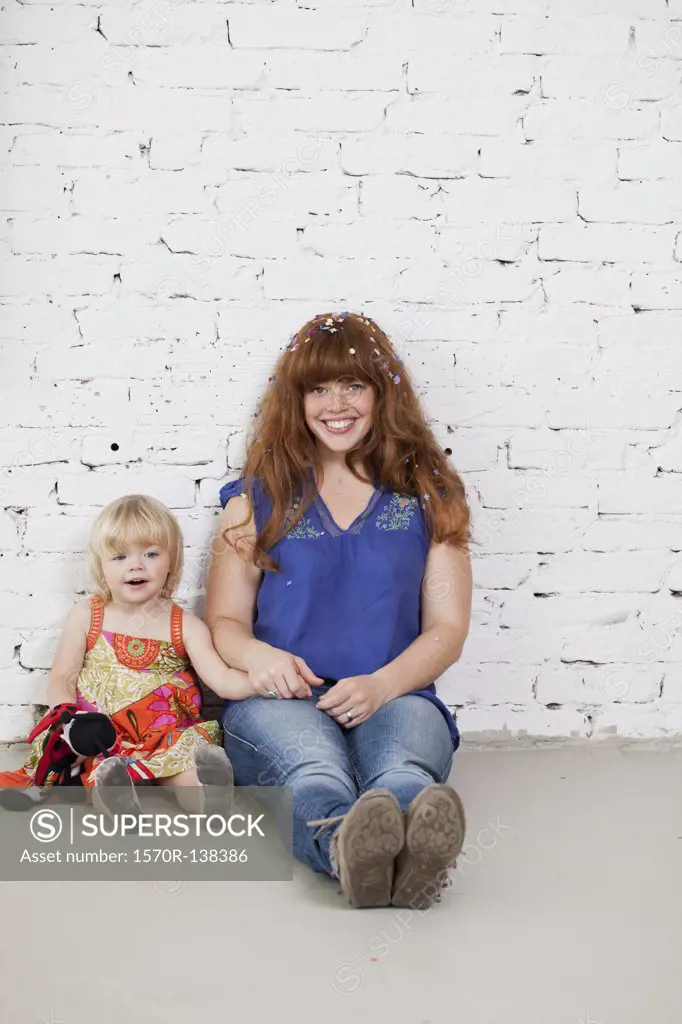 A mother and her young daughter sitting side by side against a brick wall