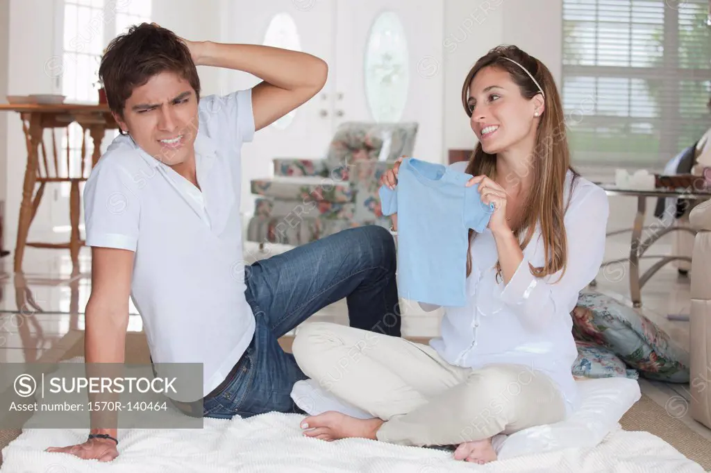 A man looking uncertain at the blue baby shirt his girlfriend is showing him