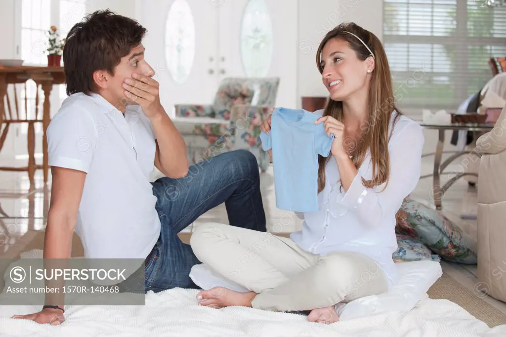 A man looking shocked at the blue baby shirt his girlfriend is showing him