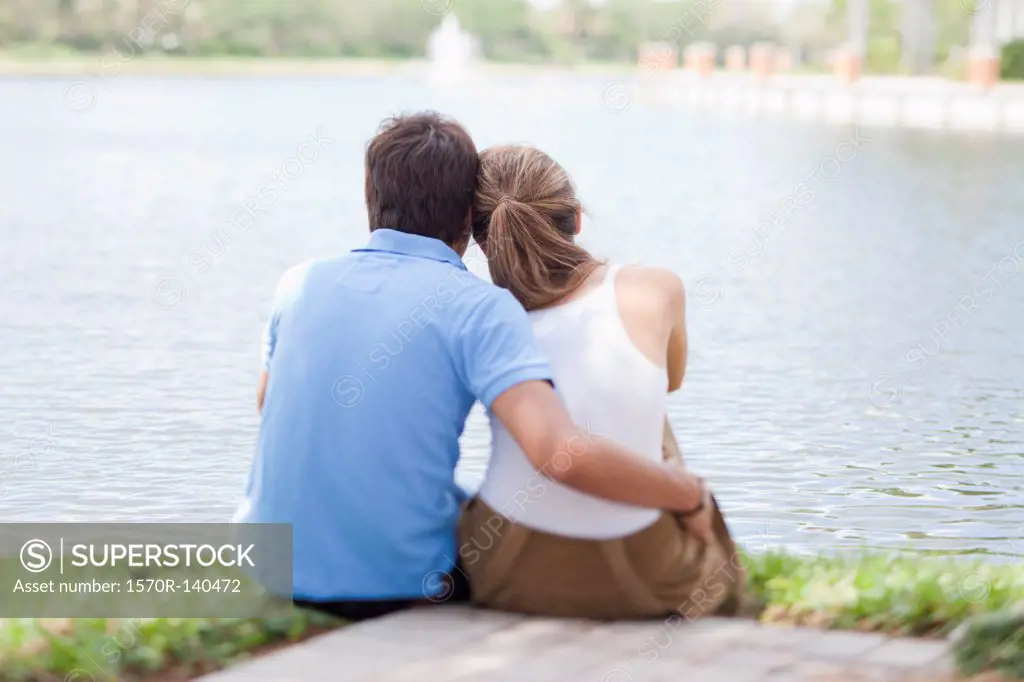 An affectionate couple sitting by a lake, rear view
