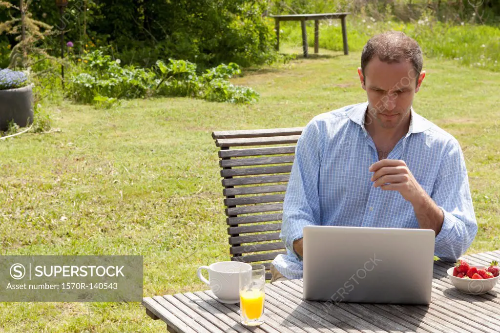 A man sitting at a table in his backyard having breakfast and using a laptop