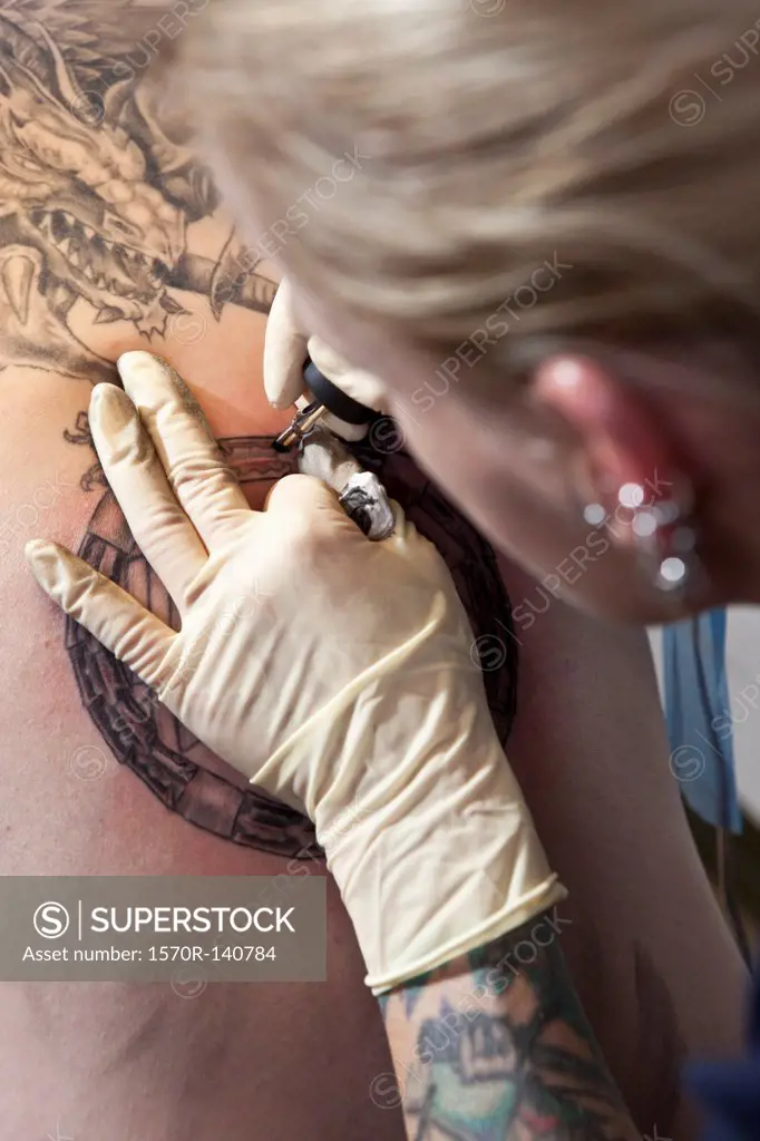 A female tattoo artist tattooing a design on a man's back, over the shoulder view