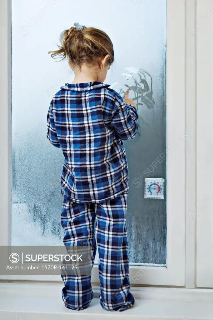A girl dressed in pajamas and drawing in condensation on glass
