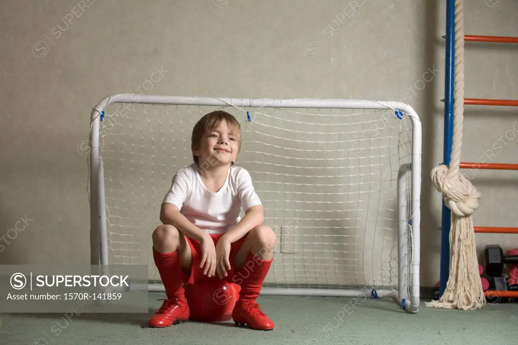 A young boy sitting on a soccer ball in front of a soccer goal