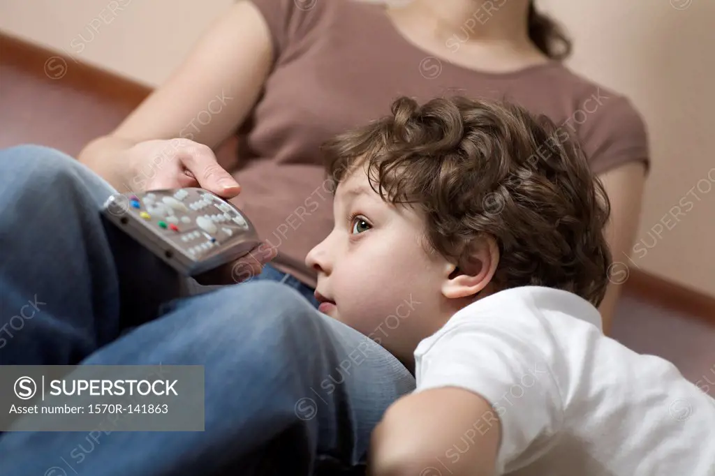 A young boy resting his head on his mom's leg, watching TV