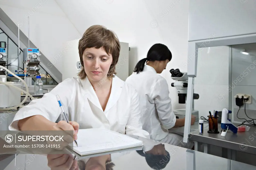 A lab technician writing notes while another technician works in background