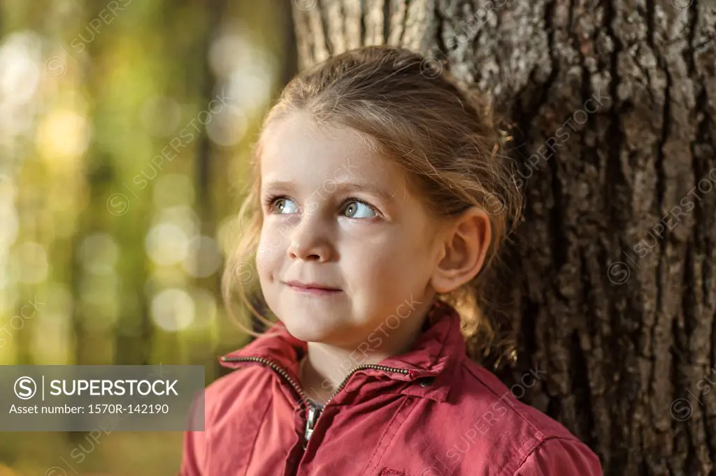 A young girl looking away curiously while leaning against a tree trunk