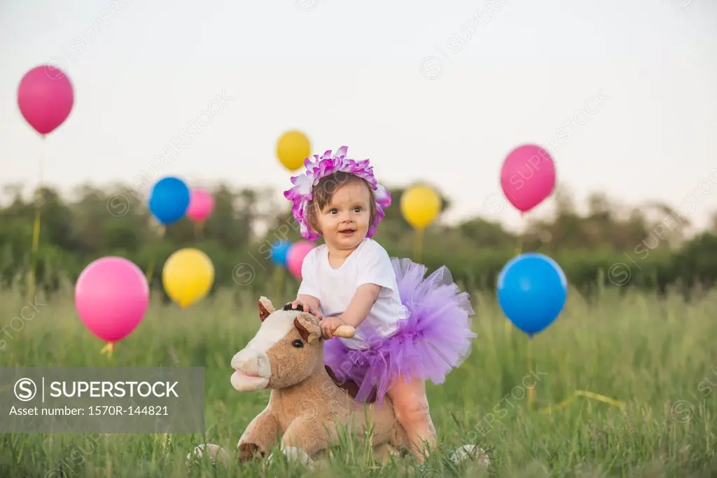A baby girl with tutu on a rocking horse in a field with balloons