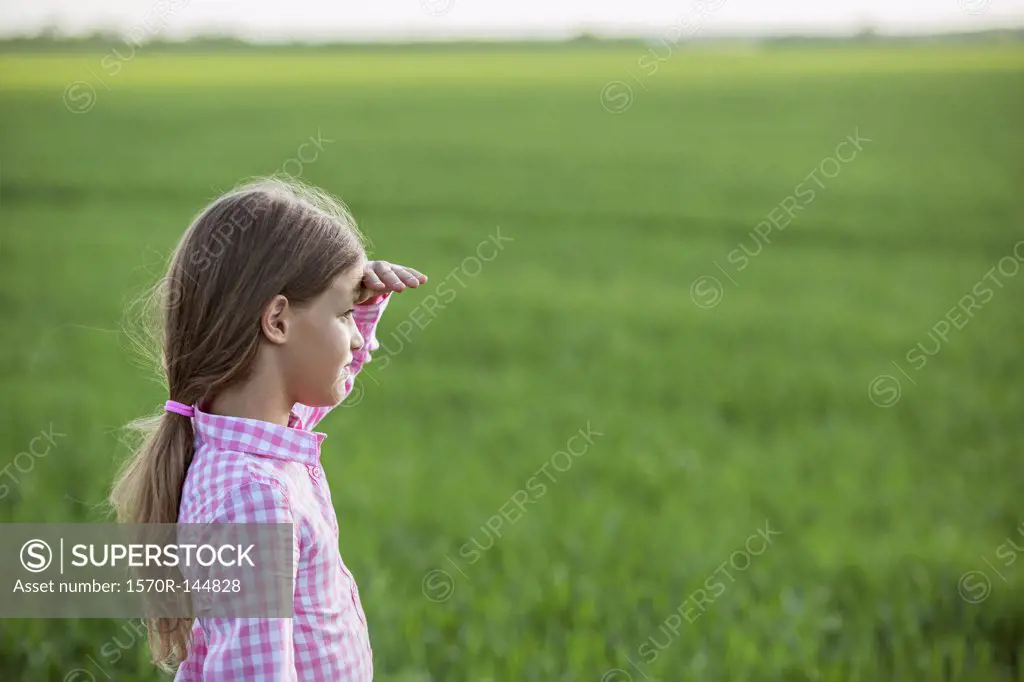 A young girl standing in a field, shielding her eyes and looking at view