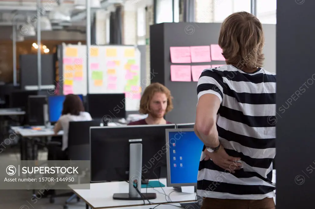 Man standing and stretching by computer desk, colleagues working in background