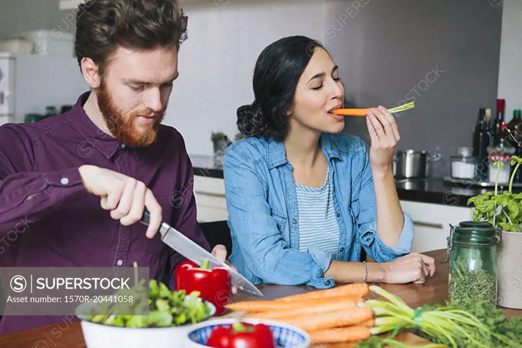 Young man chopping red bell pepper beside woman eating carrot at kitchen table