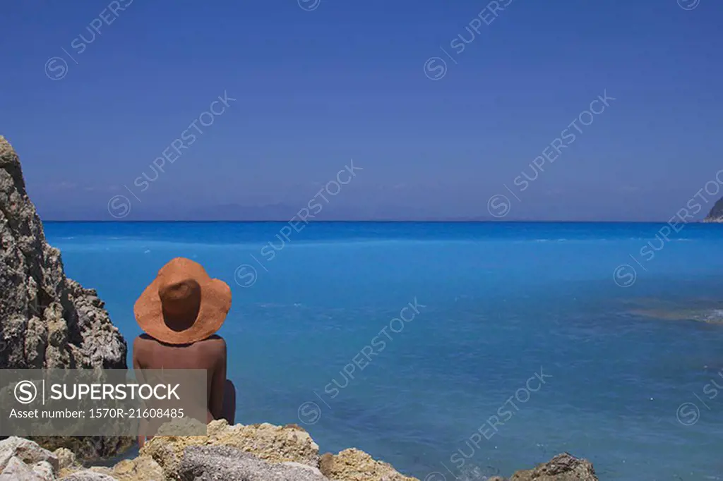 Rear view of woman relaxing on rocky sea shore against clear blue sky