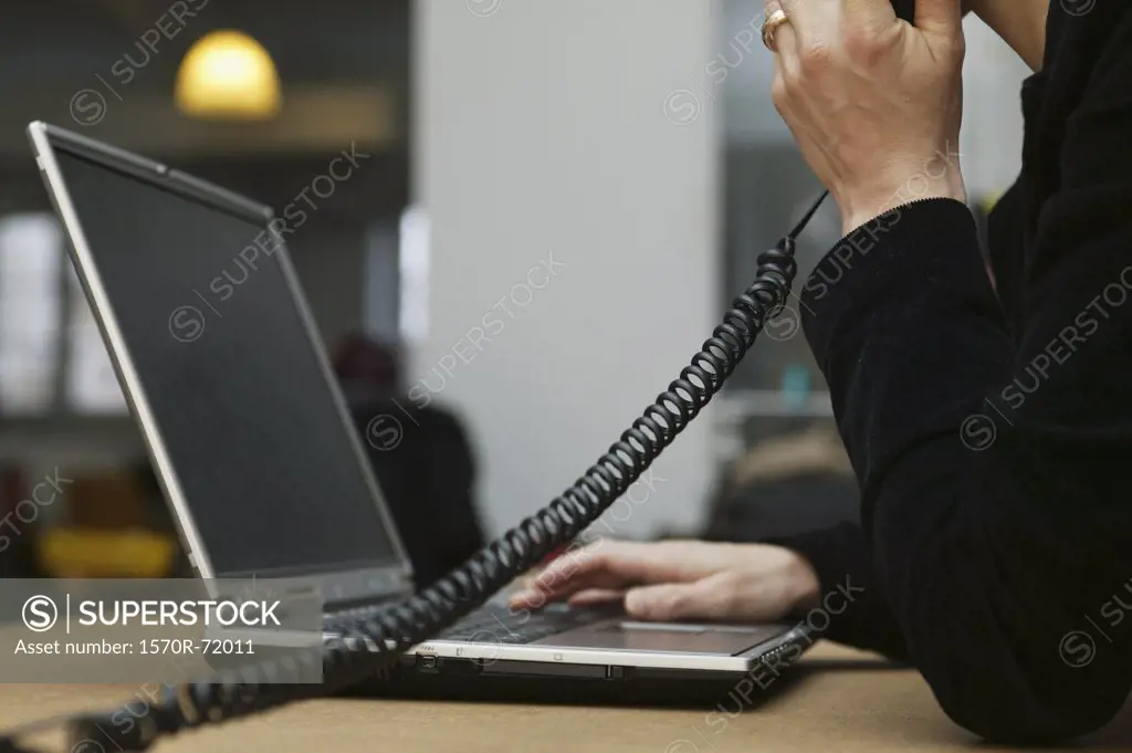 A woman sitting at a desk, using a laptop while talking on a telephone