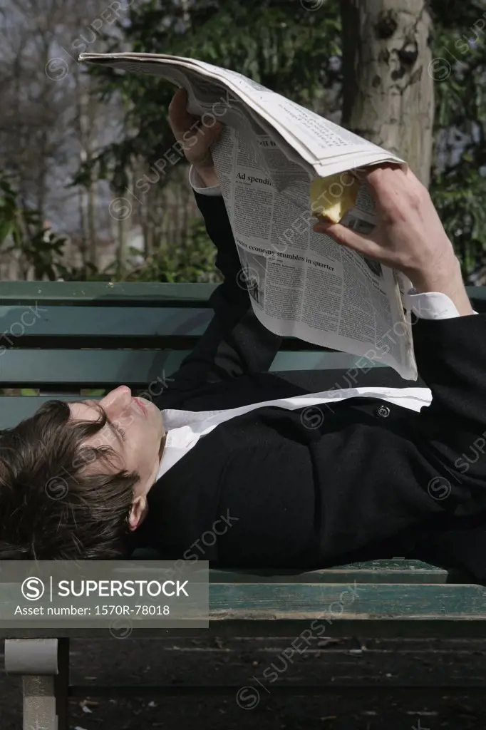 A businessman lying down on a park bench reading a newspaper and eating an apple