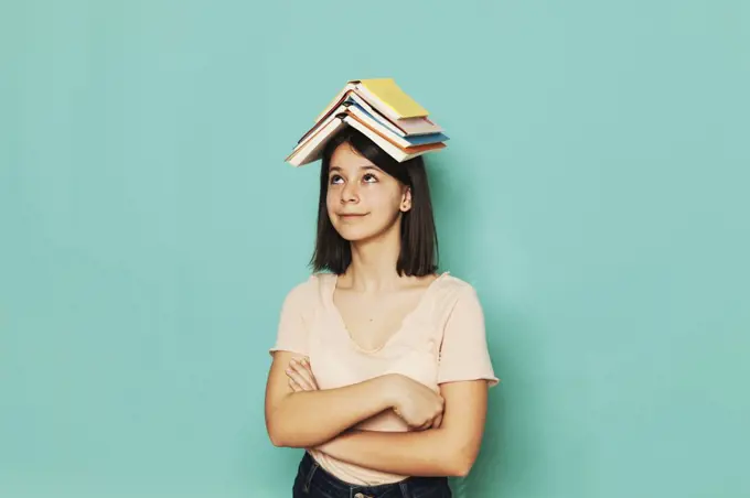 Smiling girl balancing book with arms crossed against turquoise background