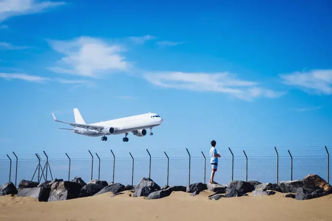 Boy on beach watching airplane flying low in blue sky near Lanzarote Airport, Spain