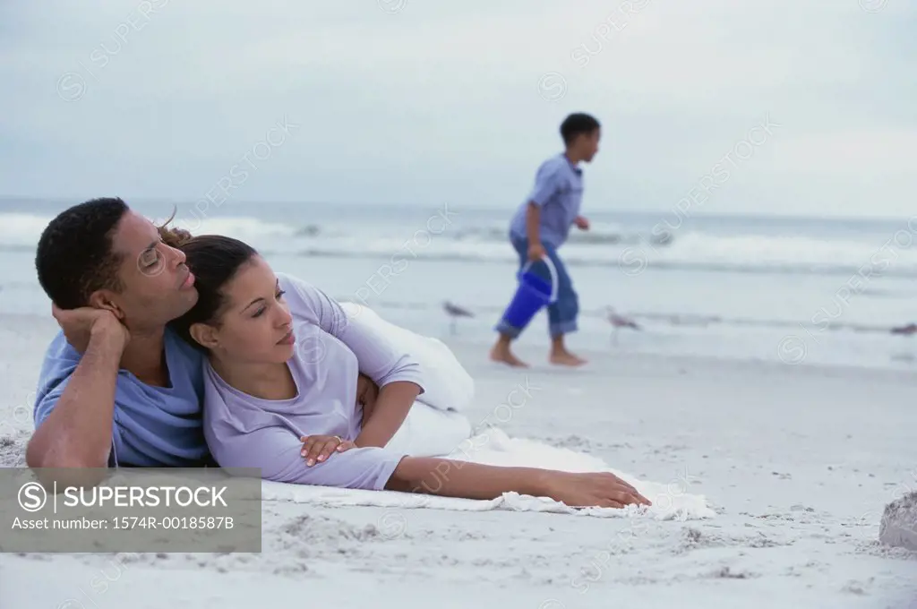 Parents lying on the beach with their son walking in the background