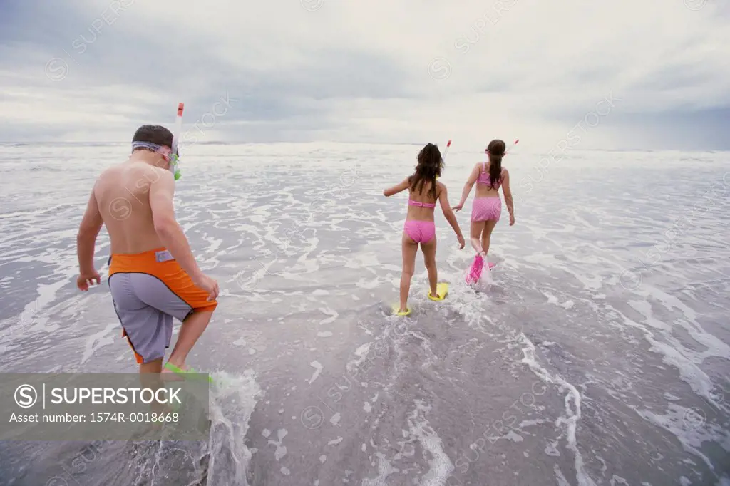 Rear view of a boy and two girls walking with snorkel masks and flippers on the beach