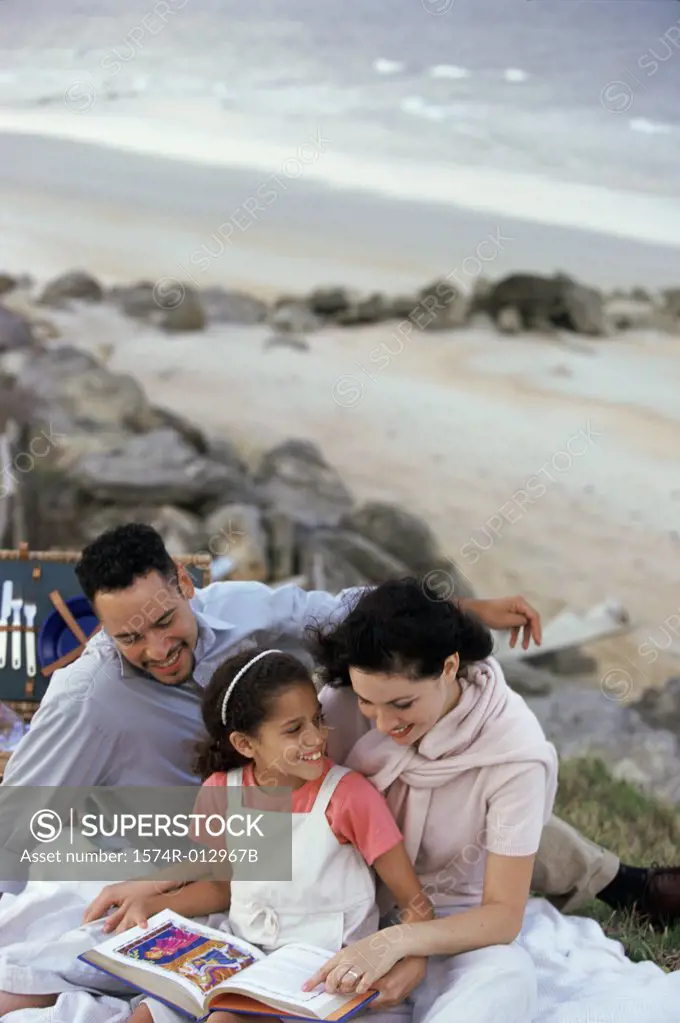 Young couple and their daughter reading a book on the beach
