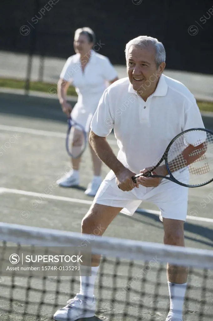 Senior man playing tennis with a senior woman in the background