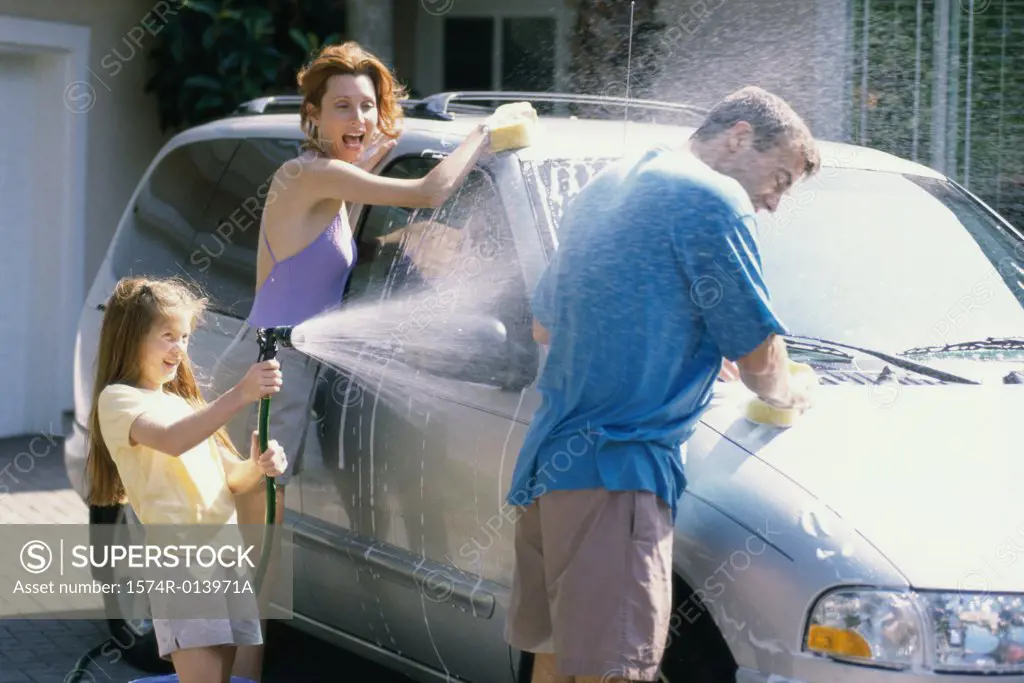 Daughter spraying water on her father with her mother washing the car