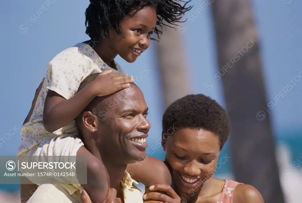 Close-up of a daughter riding on her father's shoulders on the beach with her mother standing beside them