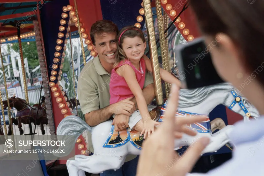 Rear view of a woman taking a photograph of her husband and daughter sitting on a carousel horse