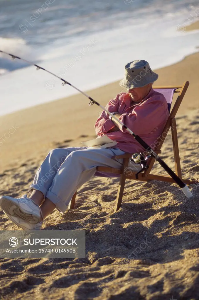 Senior man sitting on the beach in a deck chair with a fishing rod -  SuperStock