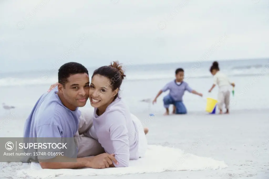 Portrait of parents lying on the beach with their son and daughter playing behind them
