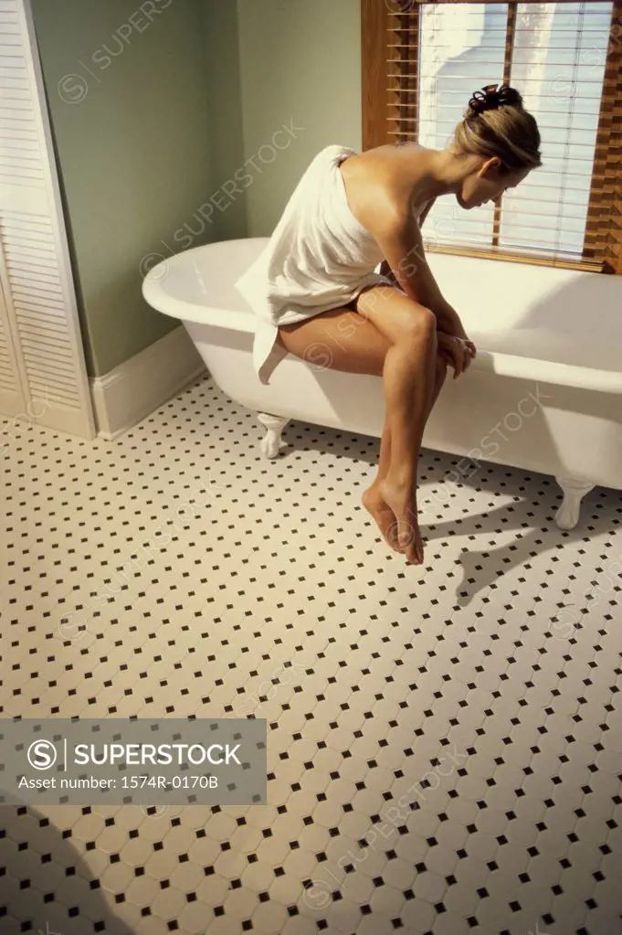 Young woman sitting in a towel on the edge of a bathtub