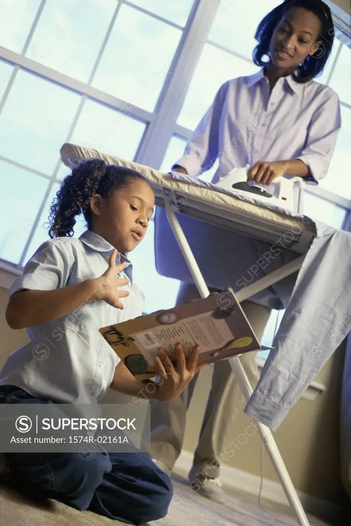 Mother ironing clothes with her daughter sitting on floor reading