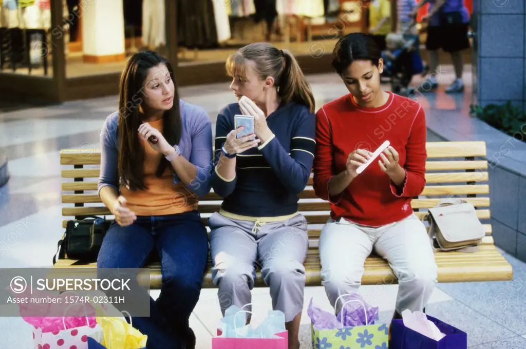 Three teenage girls sitting on a bench in a shopping mall applying make-up