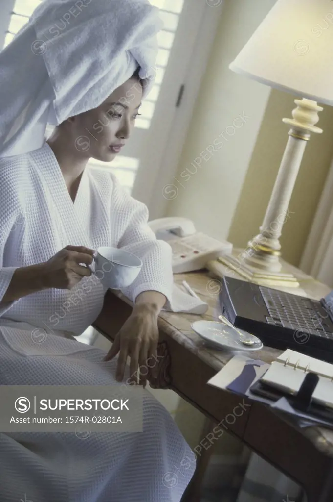 Young woman wearing a bathrobe holding a cup of coffee and looking at a laptop in a hotel room