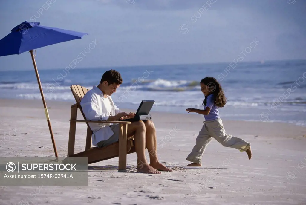 Father working on a laptop with his daughter running on the beach