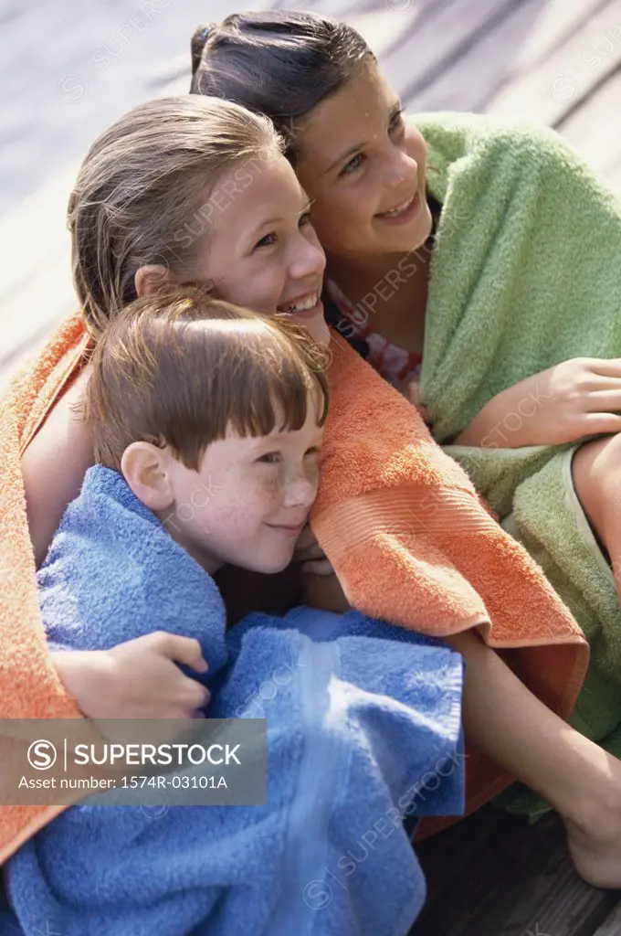 Two girls and a boy wrapped in towels sitting on a dock