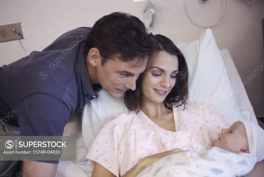 Mother lying in bed holding her baby boy with father standing beside them