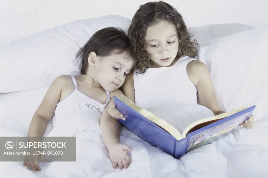 Two sisters lying in bed together reading a book