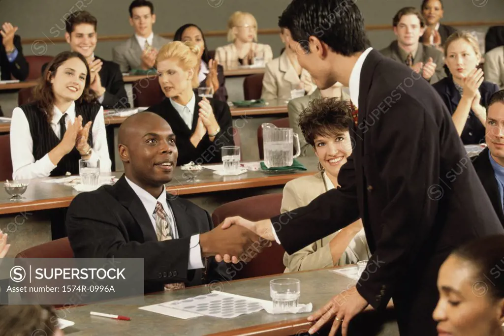 Businessmen shaking hands with people clapping in the background