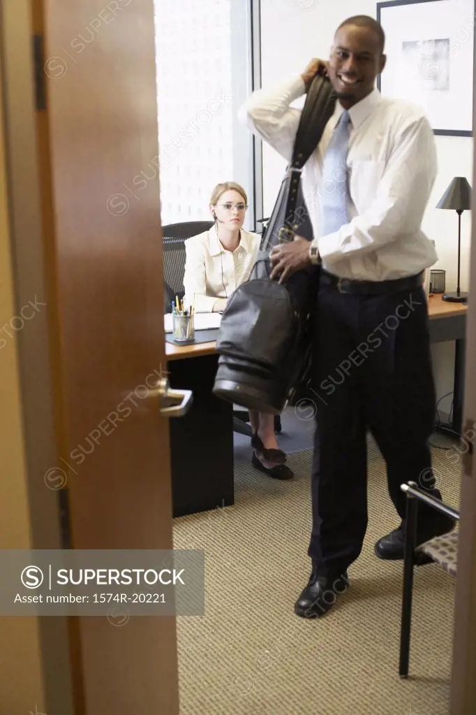 Portrait of a businessman carrying his bag with a businesswoman sitting in the background in an office
