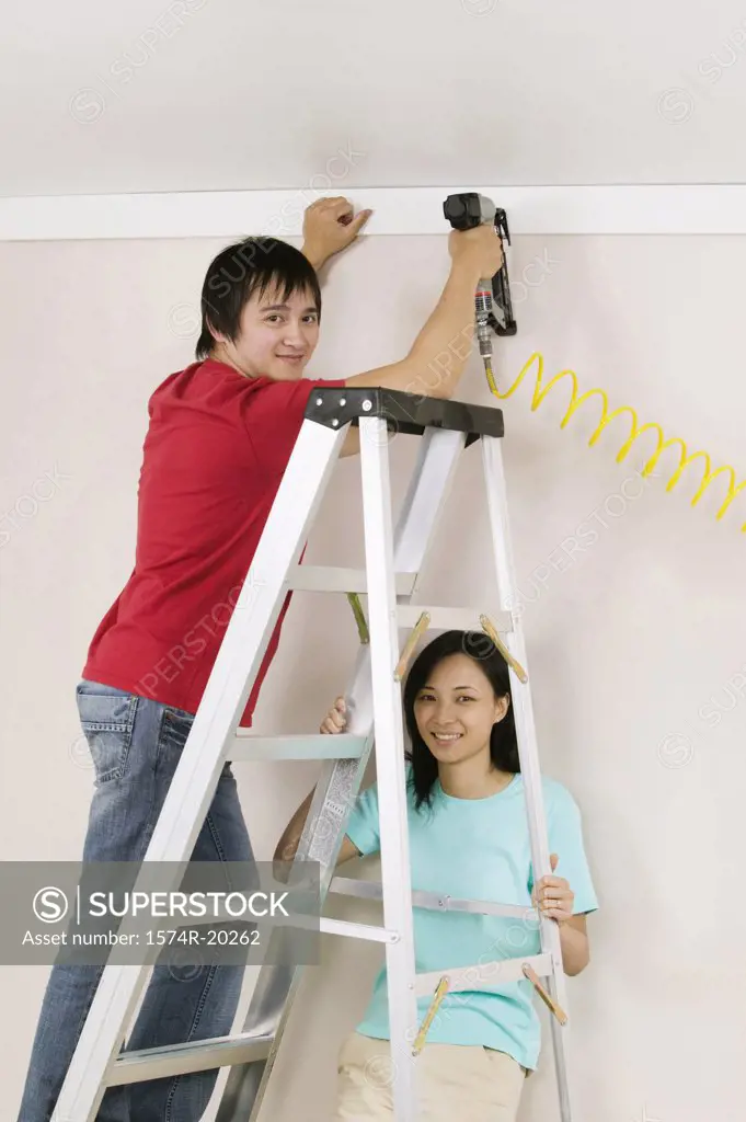 Portrait of a young man using a drill on a wall with a young woman holding a ladder
