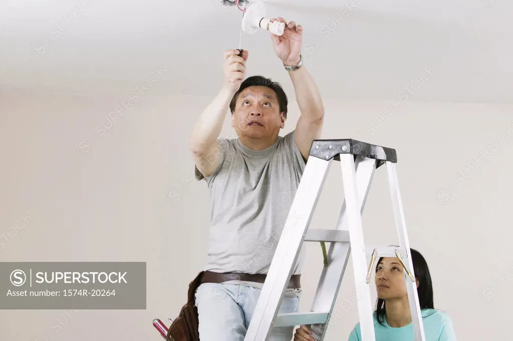 Low angle view of a mid adult man fixing a light fixture with a young woman holding a ladder