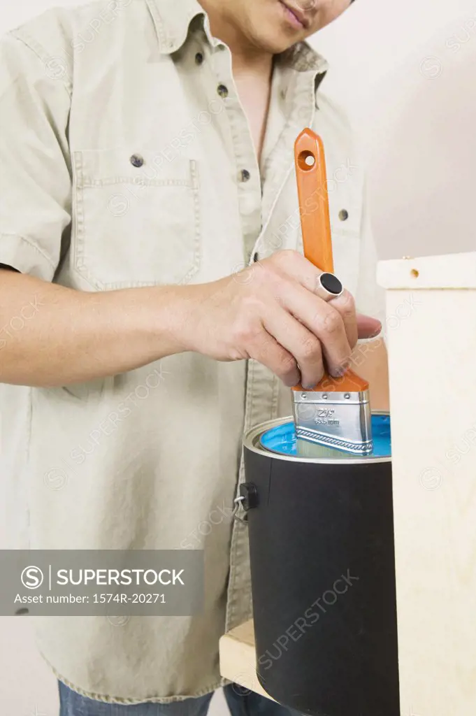 Mid section view of a young man dipping a paintbrush into a paint can