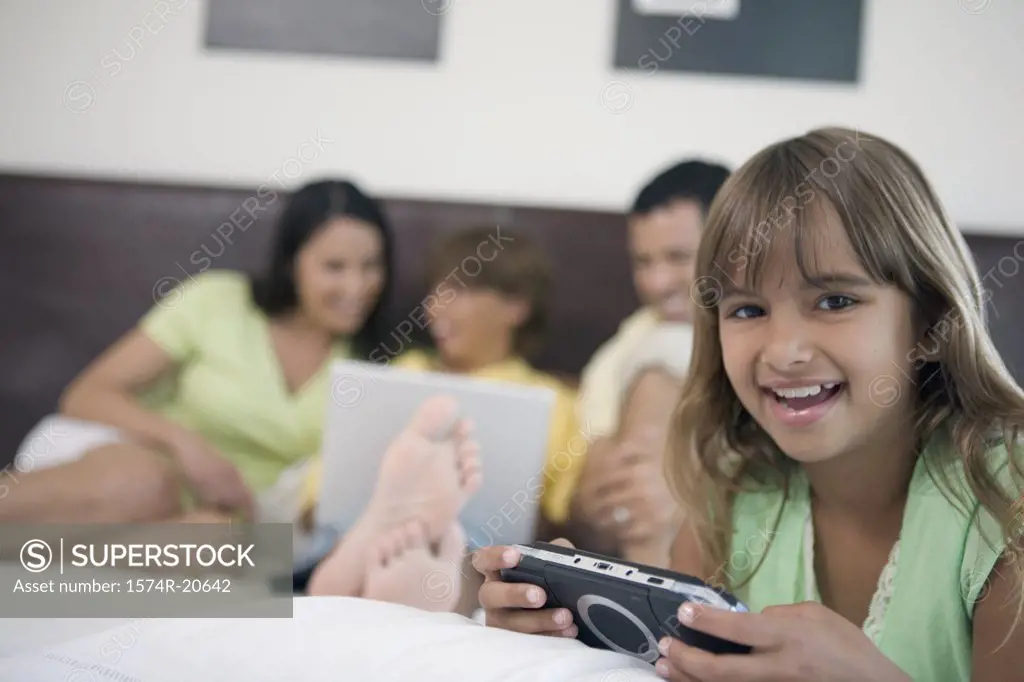 Portrait of a girl holding a handheld video game with her parents and brother looking at a laptop in the background