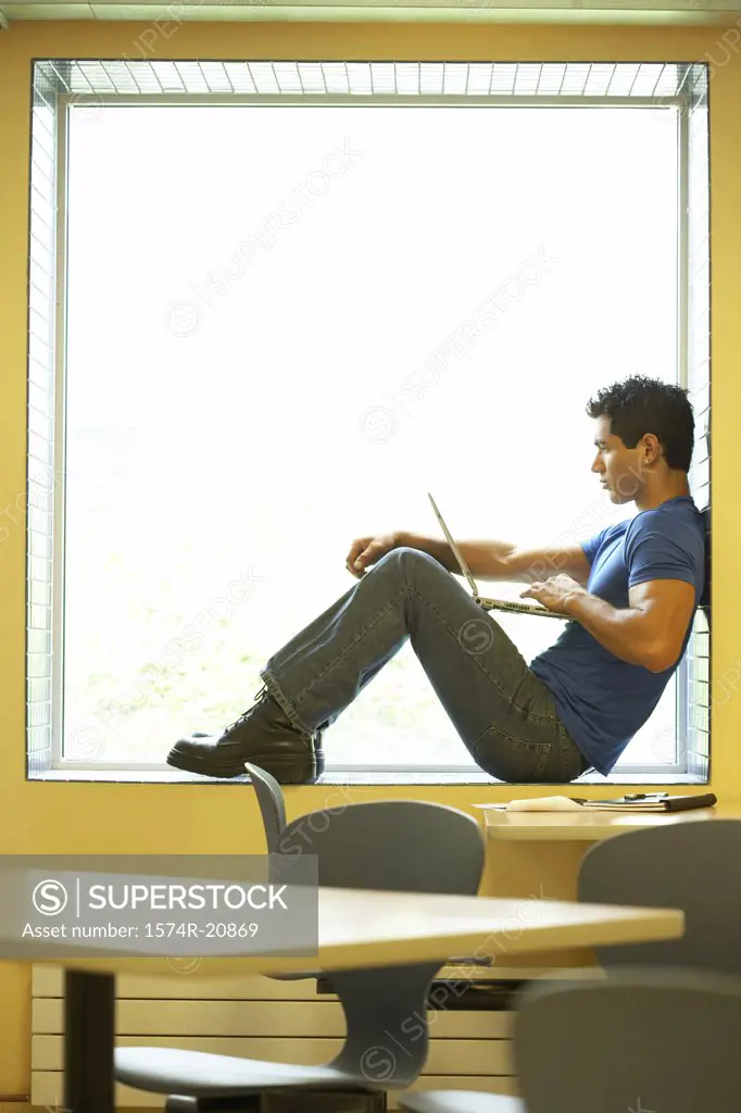 Side profile of a college student sitting on a window ledge and using a laptop