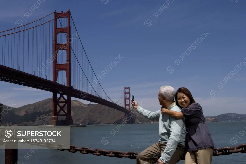 Mature man taking a picture of a suspension bridge with a mature woman embracing him from behind, Golden Gate Bridge, San Francisco, California, USA