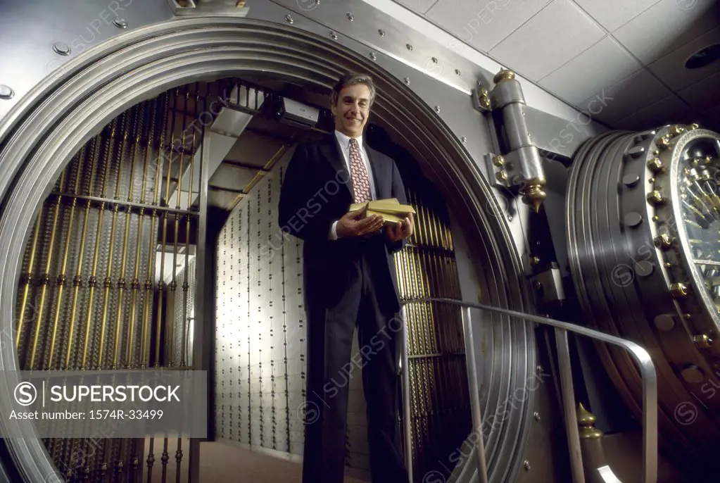 Low angle view of a businessman standing in front of a vaulted door and holding gold bars