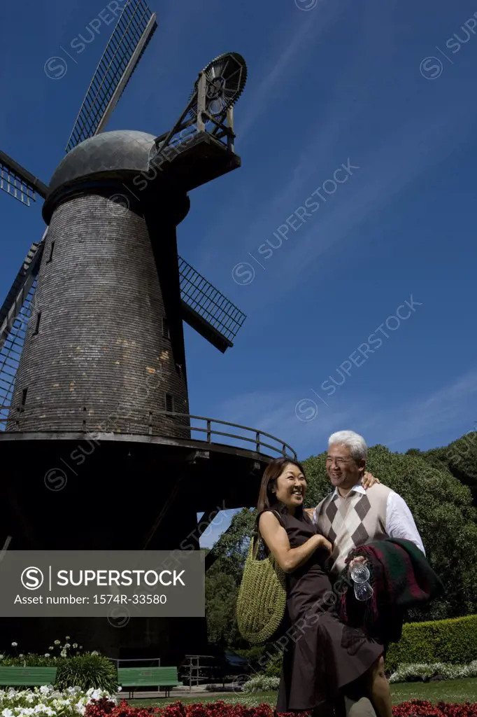 Mature couple standing in front of a traditional windmill in a park and smiling, Golden Gate Park, San Francisco, California, USA