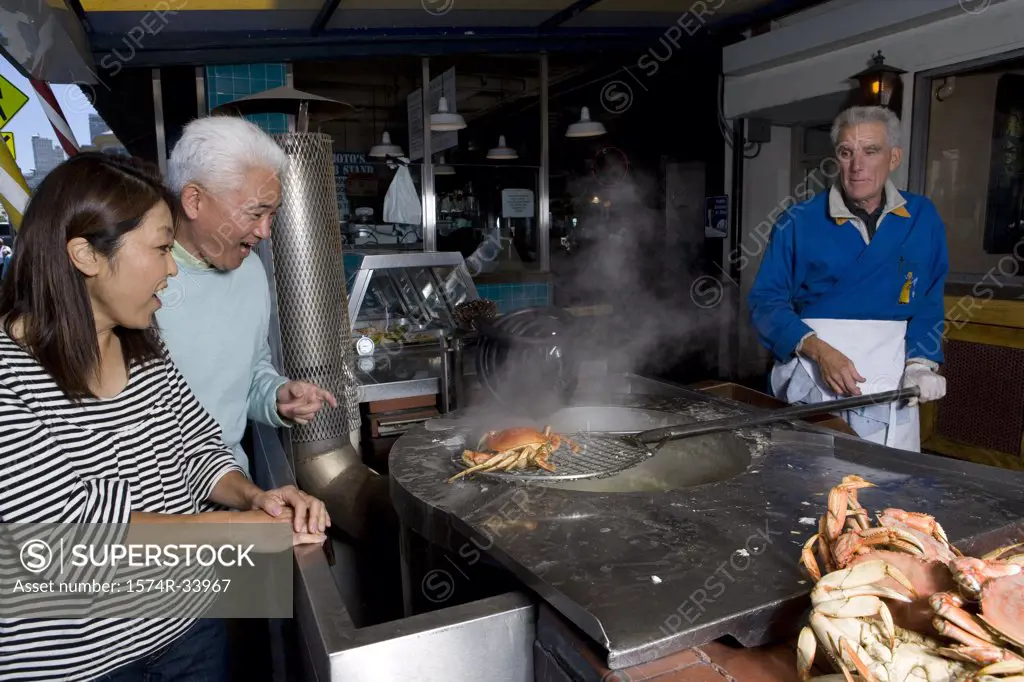 Mature couple standing and looking at crabs being cooked by a mature man in a restaurant, Fisherman's Wharf, San Francisco, California, USA