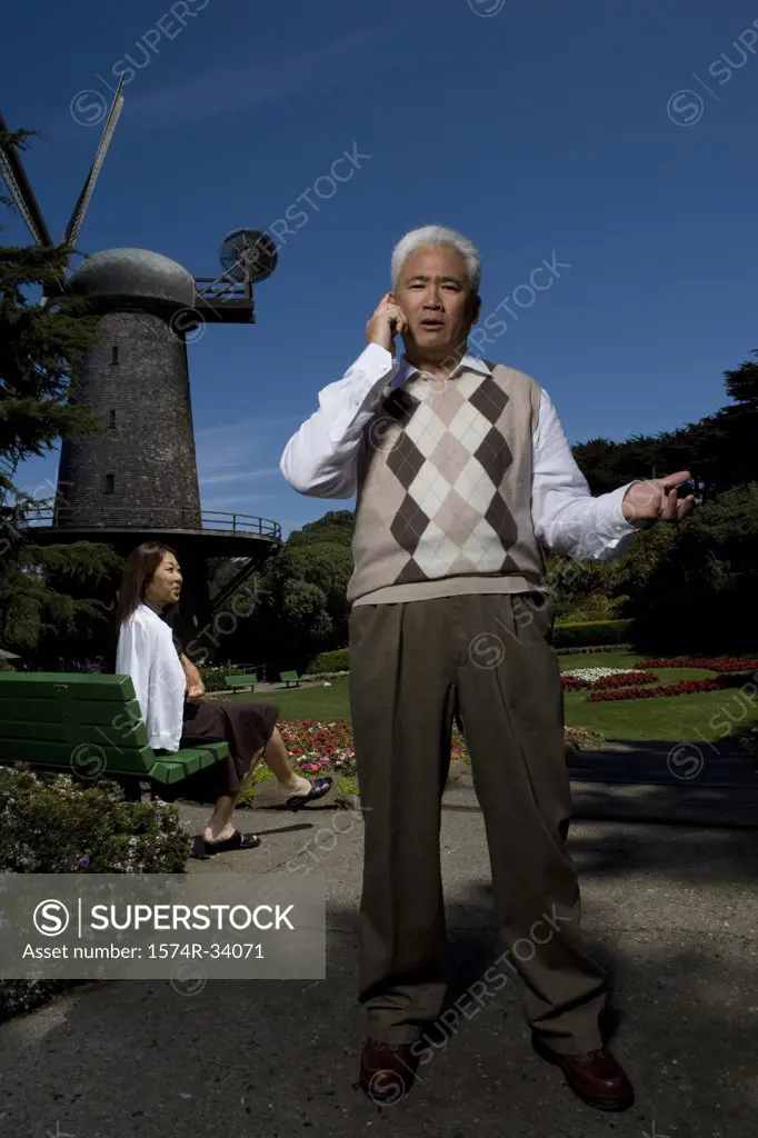 Mature man talking on a mobile phone with a mature woman sitting on a bench in the background, Golden Gate Park, San Francisco, California, USA