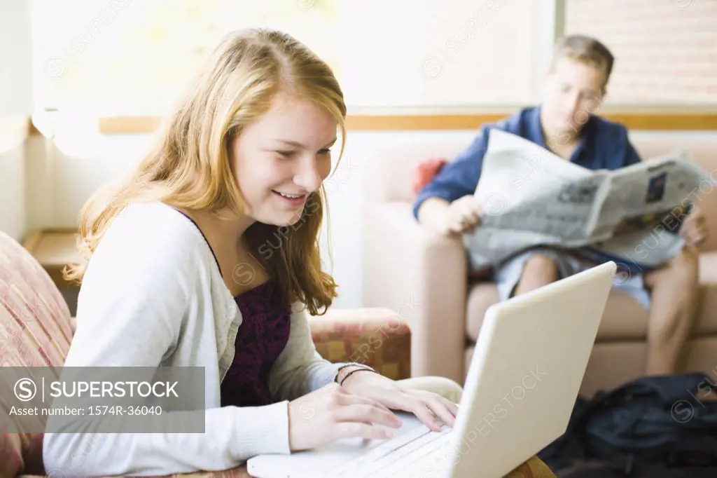 Teenage girl working on a laptop and a teenage boy reading a newspaper beside her